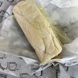 Fat Doobie - A sandwich or burrito with a soft, white bread or tortilla wrap is shown, partially unwrapped and resting on branded paper with Fat Shack logos. The food appears to be a takeout item, with some visible grease or oil on the wrapping.