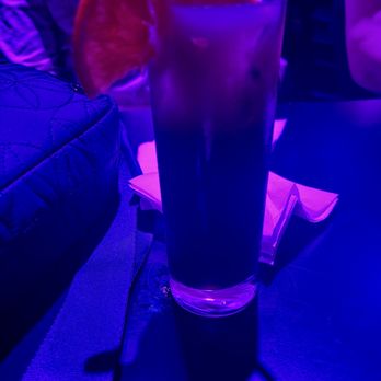 Comedy Connection - Yummy drinks - A tall, transparent glass contains a dark beverage, possibly a cocktail, with a slice of grapefruit on the rim as garnish. The table surface is dark, and there are some napkins and a quilted bag nearby. The background features purple and blue lighting, indicating a nightclub or bar environment. - Comedy Clubs Near Me - East Providence, Rhode Island