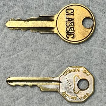 Premier Mobile Locksmith - 1940 Buick Special ignition/door & trunk key (bottom) and fuel door key (top) - Two metal keys are placed on a gray textured surface. The top key has a round head with the word "CLASSIC" engraved on it. The bottom key has a hexagonal head with detailed engravings, including "ABLOY" and other markings. Both keys are positioned horizontally, with the key blades pointing to the left. - Keys & Locksmiths Near Me - Tustin, California