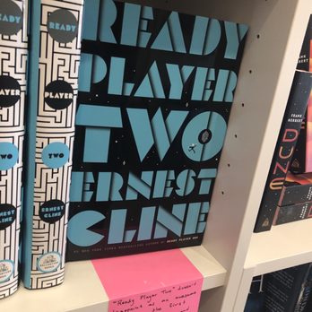 Beastly Books - A hardcover copy of 'Ready Player Two' by Ernest Cline is placed on a white bookshelf, with other books nearby. The book cover features large, stylized blue and black text. To the left, there are vertically stacked books with a black and white geometric design and blue circles. A pink note with handwritten text is attached to the shelf below the book, quoting a positive review of 'Ready Player Two.' The setting appears to be a bookstore or library, specifically an indie bookstore specializing in speculative fiction. - Bookstores Near Me - Santa Fe, New Mexico