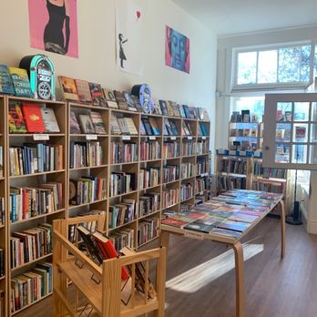 Sleepy Cat Books - The image shows the interior of a bookstore with multiple wooden bookshelves filled with a variety of books. A table in the center displays more books, and a small wooden rack holds additional titles. The wall features three pieces of artwork, including a portrait of a woman, a silhouette of a girl, and a blue-toned face. Natural light streams in through large windows, illuminating the space and creating a cozy atmosphere. - Bookstores Near Me - Berkeley, California
