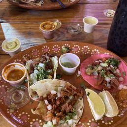 Street tacos and a quesadilla. - A plate of Mexican street tacos, a quesadilla, and various condiments and lemon wedges are displayed on a wooden table at a restaurant.