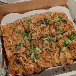 A slice of pizza in a cardboard box is topped with fried onions, chopped green onions, and a drizzle of sauce. The pizza appears to have a crispy crust and is garnished with fresh green onions. The box is placed on a table with a paper plate and a plastic utensil visible in the background.