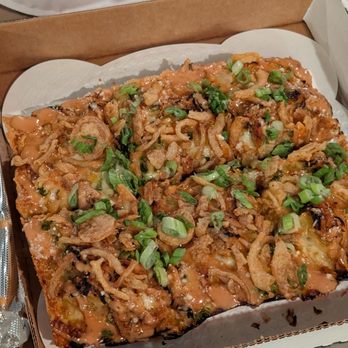 Futuro - A slice of pizza in a cardboard box is topped with fried onions, chopped green onions, and a drizzle of sauce. The pizza appears to have a crispy crust and is garnished with fresh green onions. The box is placed on a table with a paper plate and a plastic utensil visible in the background. - Pizza Near Me - Indianapolis, Indiana