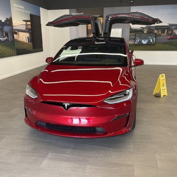 Tesla - Model X - A red Tesla Model X with its falcon wing doors open is displayed in a showroom with informational posters on the walls. - Car Dealers Near Me - Bartlett, Tennessee