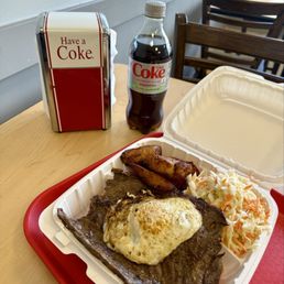 Bistec a Lo Pobre - A takeout meal served in a white plastic container on a red tray includes a steak topped with a fried egg, two pieces of fried plantains, and a side of coleslaw. On the table, there is a bottle of Diet Coke and a napkin dispenser with a sign that says "Have a Coke." The setting appears to be inside a casual restaurant or diner with wooden chairs and a light-colored wall in the background.