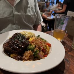 Asian Short Rib - A white round plate contains Asian short ribs with a dark glaze, served alongside rice mixed with broccoli, red bell peppers, and onions, and a small portion of pasta with a light sauce. The plate is on a wooden table in a restaurant with visible background patrons and a glass of beer nearby. The setting appears casual with a lively atmosphere.