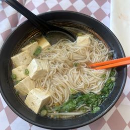 Nam Daeng !!! So good!!!! - A black bowl of noodle soup features tofu cubes, chopped green onions, cilantro, and cooked noodles submerged in broth. A black spoon rests inside the bowl, and orange chopsticks are placed on the side. The bowl is on a checkered tablecloth with a pink and white pattern, and part of a plate is visible in the background.