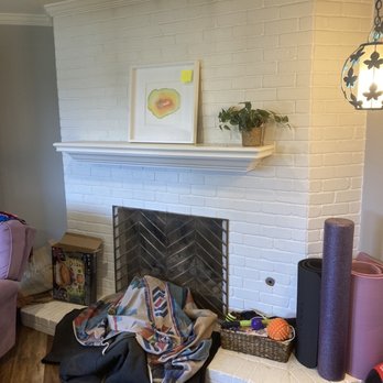 Rodriguez Tile Work - Before photo of painted brick - A white brick fireplace with a mantel features a framed artwork and a potted plant. Items like a blanket, exercise equipment, and toys are nearby. - Flooring Near Me