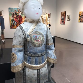 Blue Rain Gallery - Blown glass - A ceramic sculpture of a young girl dressed in traditional Asian attire, featuring detailed embroidery and metallic accents, is displayed on a black pedestal in an art gallery. The gallery has white walls decorated with colorful paintings, and visitors are seen observing the artwork. - Art Galleries Near Me - Santa Fe, New Mexico
