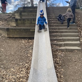 Alms Park - Children are engaging in play on a concrete slide at Alms Park, Cincinnati, OH. One child is sliding down, wearing a blue jacket and a hat with a cartoon character. Another child is climbing the stairs, and a girl at the top is preparing to slide. Adults are present, with one man looking at his phone and another woman standing nearby. The park features leaf-covered ground, wooden retaining walls, and leafless trees under a partly cloudy sky. - Parks Near Me - Cincinnati, Ohio