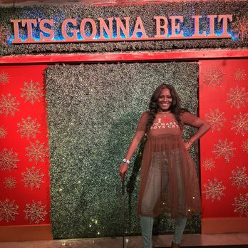 Shipwrecked - I liked the step n repeat they had for the holidays - A woman poses in front of a festive backdrop decorated with snowflakes and greenery, with a sign that says "IT'S GONNA BE LIT." She is smiling and dressed stylishly, holding a cane. - Mini Golf Near Me - Brooklyn, New York