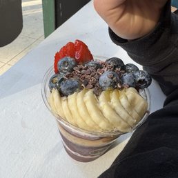 Acai Bowl - 12oz - A fruit parfait in a clear plastic cup features layers of chocolate, fruit, and possibly yogurt or mousse. The top is garnished with sliced bananas, blueberries, a strawberry, and chocolate shavings. The cup is placed on a white table near a window, with a person's hand and arm visible in the background, wearing a black sleeve.
