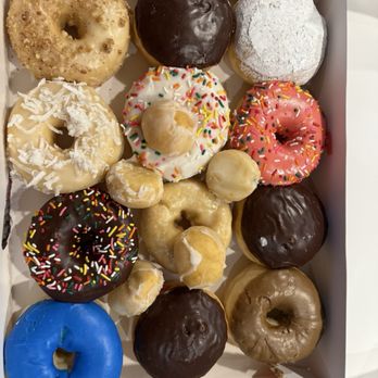 Cronuts Donuts - Assorted dozen - A box contains a variety of donuts with different toppings, including chocolate, powdered sugar, sprinkles, and coconut, arranged on a gray countertop. - Donuts Near Me - Rogers, Arkansas
