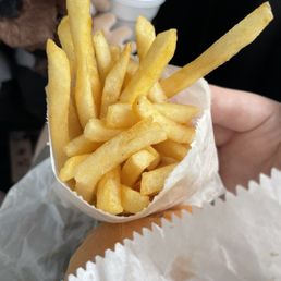 Val's small fry - A serving of French fries in a paper cone is held by a person, with a background showing a hand holding a drink and a dog.