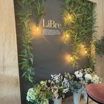 LiBre Dessert Cafe - The image shows a decorative wall at LiBre Dessert Cafe with a dark background, adorned with green fern plants and hanging Edison-style light bulbs emitting warm light. At the base, there are four white pots filled with white and green floral arrangements. The wall includes the cafe's name, LiBre, and a brief description of its offerings. The setting appears cozy and designed for aesthetic appeal. - Coffee & Tea Near Me - Hamburg, New Jersey