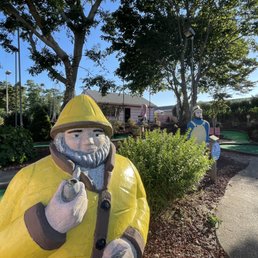 Statues of gnomes dressed in yellow and blue are situated along a winding pathway of a mini golf course. The scene includes trees, bushes, and a small building in the background, with outdoor lighting and a clear sky overhead.