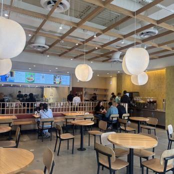 CAVA - The image shows the interior of a cafe or casual dining restaurant with a contemporary design. Wooden tables and chairs are arranged throughout the space, with some customers seated and eating. The ceiling has a wooden grid structure with hanging pendant lights. In the background, a counter with menu screens and staff preparing food is visible, along with a yellow accent wall and shelving. The overall atmosphere is bright and welcoming. - Mediterranean Near Me - Arlington, Texas