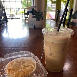 A plastic cup of iced coffee with a black straw is placed on a wooden table inside a cafe. Next to it is a cupcake in a clear plastic container. The cafe has large windows allowing natural light, with a view of the outside, and green doors with wreath decorations. A small potted plant and a spray bottle are also on the table.