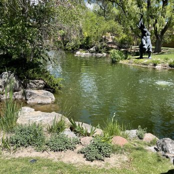 Nedra Matteucci Galleries - The garden in back of the gallery - A pond with green water is bordered by rocks, grass, and various plants, with trees overhead and a sculpture of an elephant on the right. - Art Galleries Near Me - Santa Fe, New Mexico
