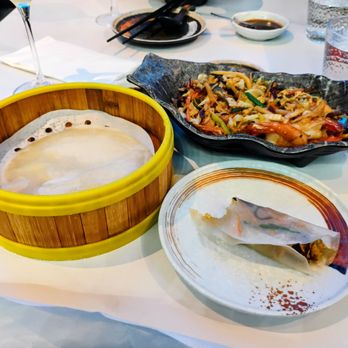 Juicy Dumplings - Mu Shu Shrimp - The image shows a restaurant table with various Asian dishes. A bamboo steamer contains a steamed dish covered with a white cloth. Next to it, a black ceramic dish holds stir-fried vegetables with shrimp, including cabbage, carrots, and green onions. A spring roll is placed on a white plate with a decorative brown rim. In the background, there are glasses, soy sauce, chopsticks, and other tableware, indicating a dining setting with Asian cuisine. - Chinese Near Me - Euless, Texas