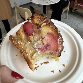 Bakey - Strawberry Broissant - A person holds a white disposable plate with a slice of cake that has fruit toppings, including strawberries and possibly peaches, garnished with almond slices and a dusting of powdered sugar. The background shows a cafe setting with a marble-topped table, a person in a white shirt sitting with a coffee, and another person standing nearby. The scene suggests a casual dining environment with customers enjoying baked goods and beverages. - Bakeries Near Me - Brookline, Massachusetts
