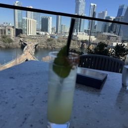 A tall glass cocktail garnished with lime and mint, with a straw, on a table with a city skyline and river in the background.
