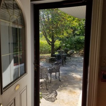 Villa Bed & Breakfast - Separate entrance - The photo captures a view from inside a building looking out through a screen door onto a patio. The patio features metal outdoor tables and chairs, with trees and greenery in the background. Sunlight filters through the leaves, casting shadows on the ground. The scene suggests a peaceful outdoor space with natural surroundings. - Hotels Near Me - Westerly, Rhode Island