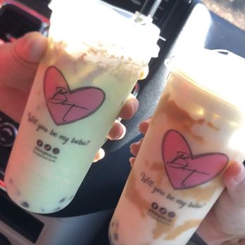 Bing Tea - Custom drink (left) honeydew you love me with coconut added and boba. Milk Thai tea with boba on the right - Two bubble tea drinks with whipped cream and caramel drizzle are held by hands inside a vehicle, with a dashboard and navigation screen visible. - Desserts Near Me - Albuquerque, New Mexico