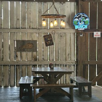 Toad's BARn & Grill - The image shows a rustic outdoor seating area with a wooden picnic table and benches inside a barn-like structure. The space features wooden walls with gaps, a wooden ceiling with exposed beams, and string lights hanging overhead. A chandelier with Edison bulbs is suspended above the table. On the wall, there is a neon Busch Light sign, a small wooden arrow sign, and a vintage American motorcycle poster. A small sign indicates no smoking. - Bars Near Me - Stanton, Kentucky
