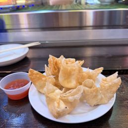 Crab Rangoon - A plate of fried wontons is placed on a dark wooden table, accompanied by a small container of red dipping sauce. The wontons are golden and crispy, arranged in a slightly overlapping manner. In the background, a restaurant window shows reflections and some outdoor elements, with a person’s arm visible behind the glass. The setting suggests a casual dining environment, likely at a Chinese or Asian cuisine restaurant.