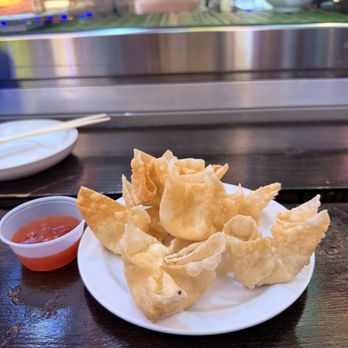 Dragon Sushi - Crab Rangoon - A plate of fried wontons is placed on a dark wooden table, accompanied by a small container of red dipping sauce. The wontons are golden and crispy, arranged in a slightly overlapping manner. In the background, a restaurant window shows reflections and some outdoor elements, with a person’s arm visible behind the glass. The setting suggests a casual dining environment, likely at a Chinese or Asian cuisine restaurant. - Sushi Bars Near Me - Albuquerque, New Mexico