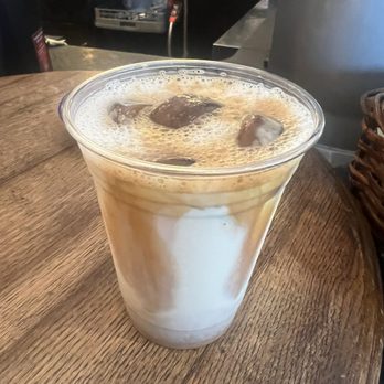 Pour Richard's Distillery - White chocolate n caramel iced coffee - A clear plastic cup filled with iced coffee, topped with ice cubes and foam, placed on a wooden table in a cafe setting. - Cafes Near Me - Devon, Pennsylvania