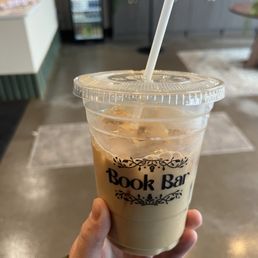 Iced latte - A person holds a plastic cup filled with iced latte, topped with ice cubes and a straw, inside a cafe named Book Bar. The cup has a black decorative logo with the text 'Book Bar.' The background shows bookshelves, a beverage fridge, and cafe furniture, indicating an indoor setting with a book-themed decor.