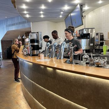 Compass Coffee - Multiple staff members are working behind a curved wooden counter in a coffee shop, with coffee machines, dispensers, and digital screens displaying menu options. A customer with a backpack is standing at the counter, and the interior features modern lighting and decor, including white tiled walls and a wood-paneled ceiling. - Coffee Roasteries Near Me - Washington, DC, District of Columbia