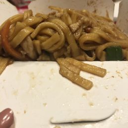 noodles in a take out box - A close-up image shows stir-fried Chinese noodles with vegetables and sauce inside a white takeout container. The noodles are partially eaten, with some sauce and food remnants visible on the container's edge. The background is blurred, indicating an indoor setting.