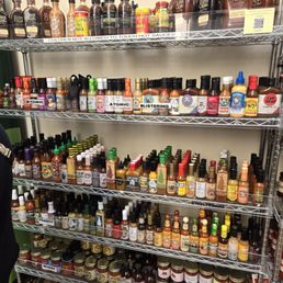 Everything hot! - A metal shelving unit holds a variety of hot sauces, seasonings, and condiments in bottles and jars. The top shelf contains larger bottles with labels, while the lower shelves feature smaller bottles and jars, organized by type and brand. The setting appears to be a retail store or storage area for hot sauce products.