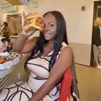 Commas Food Hall - A woman with long dark hair is seated at a restaurant, holding a large sandwich or burger near her face, with other diners visible in the background. - Bars Near Me - Silver Spring, Maryland