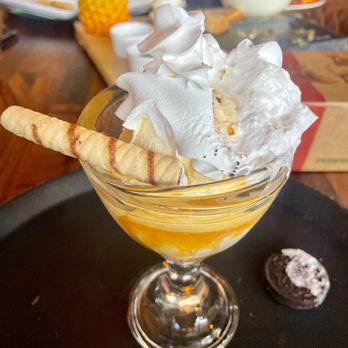 Nash & Smashed - Caramel Sundae - A dessert parfait in a clear glass features whipped cream on top, garnished with a wafer roll and a cookie placed on a black tray. The background shows a wooden table with various dishes, a pineapple decoration, and restaurant tableware, indicating a casual dining environment. - Sandwiches Near Me - Silver Spring, Maryland