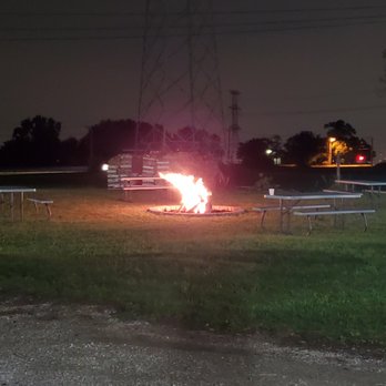 VFW - The image depicts a nighttime outdoor scene with a fire burning in a fire pit at the center. Several picnic tables are arranged around the fire, and the area appears to be a park or recreational space. In the background, there are trees, utility poles, and power lines, with some distant lights visible. The ground is covered with grass and gravel, and the overall scene is dimly lit except for the bright flames of the fire. - Veterans Organizations Near Me - Frankfort, Illinois