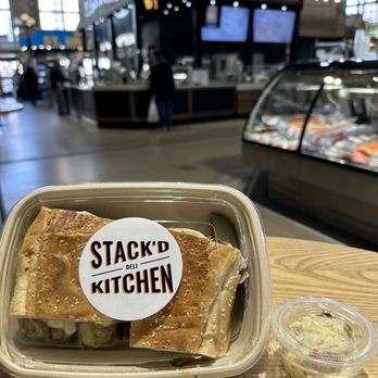 Stack'd Deli Kitchen - Roast beef mushroom melt hoagie - A sandwich with a sesame seed bun, possibly containing roast beef and mushrooms, is in a clear plastic container with a Stack'd Deli Kitchen sticker. Next to it is a small cup of coleslaw. The background shows the interior of Stack'd Deli Kitchen, a deli counter with digital menu screens, and a busy market setting with people and other food vendors. - Salad Near Me - Toronto, Ontario