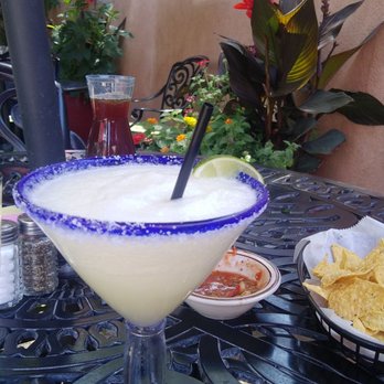 Casa De Benavidez - A margarita cocktail with a salted rim, lime wedge, and straw is on a black metal outdoor table. The table also has a bowl of chips and salsa, a pitcher of tea, and salt and pepper shakers. The setting includes potted plants, flowers, and a stucco wall with decorative elements, indicating an outdoor dining area. - Mexican Near Me - Albuquerque, New Mexico