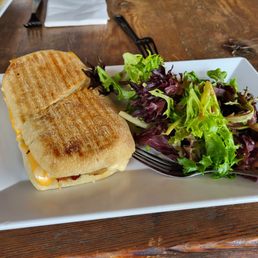 The Matthew panini - A toasted sandwich with melted cheese on a white rectangular plate, accompanied by a mixed green salad with leafy lettuce.