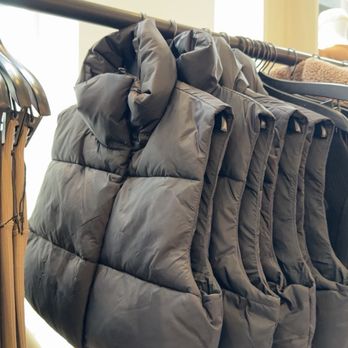 ALO - Multiple black puffer vests are hanging on a metal clothing rack inside a retail store. The vests are arranged side by side, with some overlapping. Natural light from windows illuminates the scene, highlighting the fabric's texture and the store's interior. The background includes a light-colored wall and part of a window frame. - Men's Clothing Near Me - Pasadena, California