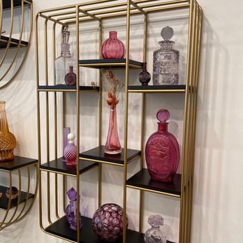 Julez Bryant - The store is filled with beautiful bottles from all over the world. Julez buys these and must have the original top.  Love this - A wall-mounted display shelf with a gold metal frame and black shelves showcases multiple glass bottles and vases in shades of pink, purple, and clear. The arrangement includes different shapes and sizes, with some featuring intricate patterns or decorative elements. The shelf is positioned against a plain, light-colored wall, and part of another similar shelf is visible on the left side of the image. - Jewelry Near Me - Encinitas, California
