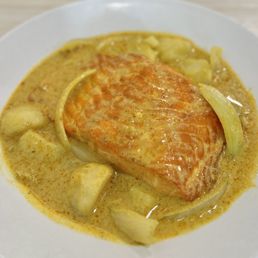 Yellow Curry - A white plate contains a piece of baked salmon topped with a yellow curry sauce. Surrounding the salmon are pieces of vegetables, possibly onions or potatoes, immersed in the curry sauce. The dish appears to be served hot, with the curry sauce covering the salmon and vegetables evenly. The presentation suggests a dish from a Thai or Asian cuisine restaurant.