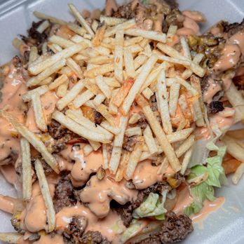 Between Two Buns - A takeout container contains a burger topped with fries, lettuce, and a pink sauce, with visible ground beef and other toppings. - Burgers Near Me - Miami, Florida