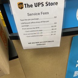 The sign is positioned on a counter at The UPS Store in Upland, CA. It details various service fees including a $2 tape fee per package, a $3 USPS/post office drop-off fee, and a $3.08 black and white print service fee, with additional charges for extra pages. Color printing costs $3.34 per page, with additional pages costing $1.32 each. The sign also states that bills over $20 are not accepted. The background includes part of the counter and some office supplies.