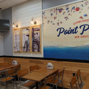 Jersey Mike's Subs - Interior - The image shows a dining area inside a restaurant with several wooden tables and matching chairs. The wall features a large mural with text and a beach scene, along with framed photographs of a bicycle by the water. There are three exposed bulb light fixtures above the artwork, and a small TV mounted on the wall. The flooring is wood-patterned, and the overall decor is casual and modern. - Fast Food Near Me - Eagan, Minnesota