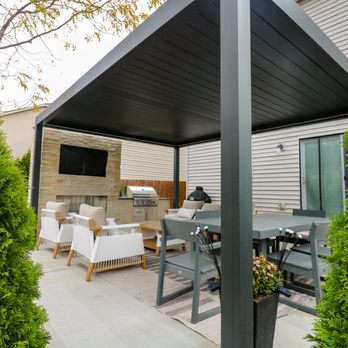 TRC Outdoor - A black pergola covering a patio area. Green arborvitae trees frame the foreground, leading the eye toward a stone fireplace. - Landscaping Near Me - Fenton, Missouri